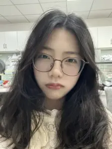 Khung hình cận mặt ghi lại cô gái tóc rối đeo kính tròn nhìn thẳng rất hiền