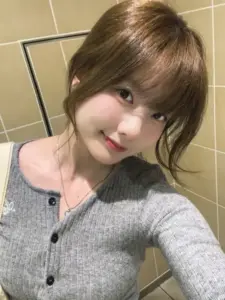 Cận mặt trước gương, cô gái Japan mặc cardigan xám mỉm cười nhẹ, tóc nâu ôm mặt