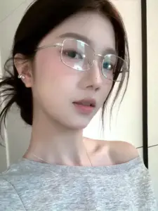 Áo lệch vai xám và kính trong khiến gương mặt cô gái sáng mềm bên cửa sổ