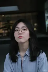 Ngoài trời tối, thiếu nữ đeo kính mặc sơ mi xanh ngước nhìn, mái tóc ngắn gọn
