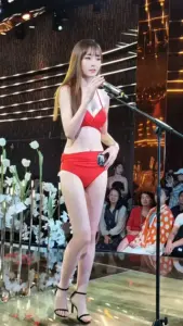 Trên sân khấu đêm, người mẫu bikini đỏ trắng mỉm cười, tay tạo dáng chữ V