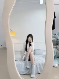 Gương bầu dục phản chiếu cô gái ngồi trên giường, điện thoại che mặt và tất dài trắng