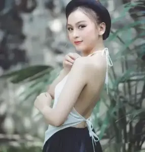 Người mẫu quay lưng giữa nền cây xanh, áo trắng hở vai, nụ cười rất nhẹ