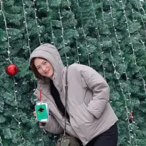 Cô gái mặc hoodie xám mỉm cười bên tường lá xanh, tay cầm điện thoại xanh mint