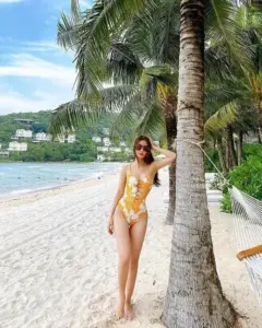 Cô gái mặc bikini vàng đứng cạnh thân dừa bên bãi cát trắng, mái tóc dài buông nhẹ