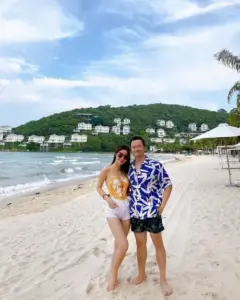 Bên bờ biển xanh, cô gái áo tắm vàng đứng cạnh người đàn ông mặc áo họa tiết