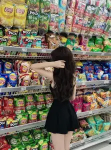 Trước kệ đồ ăn vặt, cô gái không lộ mặt diện váy đen ngắn đầy tinh nghịch