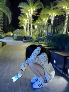 Cô nàng giấu mặt ngồi xổm bên hồ đêm, áo khoác trắng cùng điện thoại nổi bật