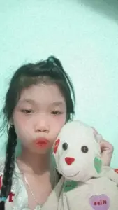 Cô gái ôm thú bông trong phòng xanh, môi chúm chím khiến ảnh có nét troll nhẹ