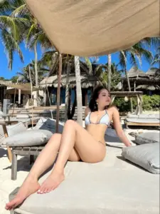 Resort biển phía sau, cô gái ngồi duỗi chân trong bikini sáng màu, kính râm rất sang