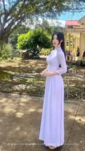 Dưới bóng cây sân trường, cô nàng mặc áo dài trắng đứng nghiêng với nụ cười nhẹ
