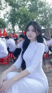 Giữa sân đông người, bạn nữ áo dài trắng ngồi nhẹ nhàng và nhìn thẳng máy ảnh