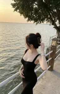 Cô gái đứng bên lan can biển, váy đen ôm dáng, tóc búi giữa hoàng hôn