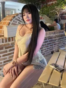 Người đẹp ngồi trước bức tường gạch, áo vàng ren, tóc đen dài thả thẳng