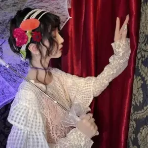 Bên rèm đỏ, cô nàng nghiêng mặt giơ chữ V, váy ren trắng và hoa cài tóc