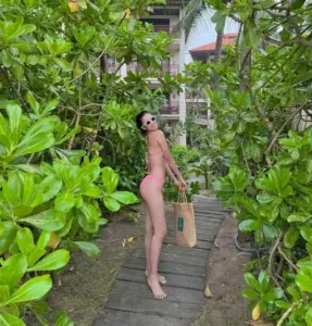 Trên lối đi xanh mát, cô gái mặc bikini hồng đứng nghiêng nhẹ giữa vườn cây rậm