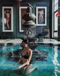Người đẹp diện bikini đen ngồi trong hồ nước xanh, ánh mắt cúi nhẹ giữa không gian sang trọng
