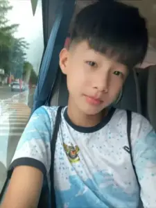 Cậu bạn mặc áo sọc xanh đứng ngoài đường, tóc đen gọn và gương mặt non trẻ dễ mến