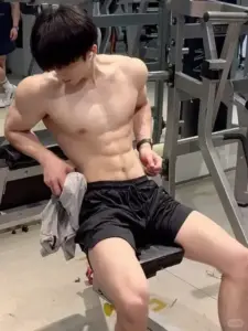 Ngồi trên ghế tập, body nam săn chắc mặc quần short đen, nhìn xuống với nét tập trung
