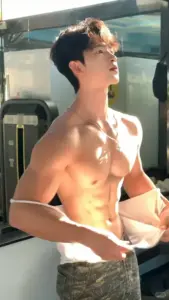 Trong phòng gym, người mẫu nam kéo áo ở eo, ngẩng mặt dưới luồng sáng ấm
