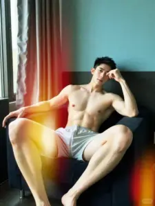 Trong góc phòng ánh màu, người mẫu nam ngồi tựa ghế, mặc quần short xám thư giãn