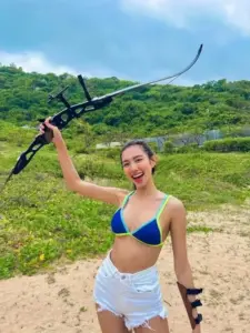 Ngoài đồi cỏ ven biển, cô gái giương cung mỉm cười, bikini xanh nổi bật