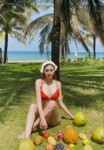 Cô gái mặc bikini đỏ ngồi dưới hàng dừa, cười nhẹ giữa nắng biển và trái dừa xanh