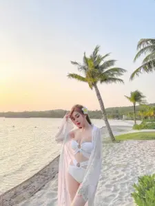Trên bãi biển chiều, diễn viên nữ mặc bikini trắng phối khăn mỏng, giơ tay tạo dáng