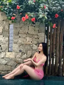 Trước tường đá hoa đỏ, cô gái mặc bikini hồng ngồi duỗi chân, nụ cười mềm mại