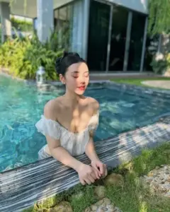 Cạnh hồ trong nắng, cô gái áo tắm trắng ngồi tựa thành gỗ, mỉm cười nghiêng mặt