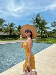 Bên hồ bơi xanh, cô gái diện đồ bơi vàng, đội nón rộng vành và nhìn xa thư thái