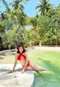 Bên hồ xanh trong cô gái mặc bikini đỏ tạo dáng ngồi, nụ cười nhẹ giữa hàng dừa