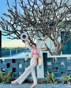 Trước bức tường xanh người mẫu đội mũ trắng, diện bikini tím và quần ren khoe dáng