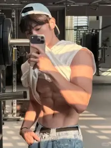 Qua gương phòng gym, mẫu nam fitness che mặt bằng điện thoại, áo hở vai, ánh nắng sọc