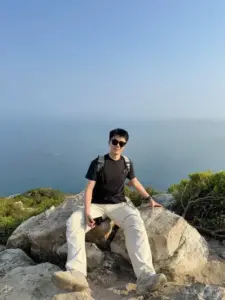 Giữa nền biển xanh mờ, chàng đeo kính ngồi trên đá với áo đen và quần sáng