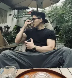 Bên bàn ngoài hiên, soái ca áo đen đeo kính nhâm nhi nước, vai rộng nổi bật
