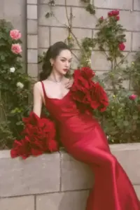 Bên khóm hoa đỏ, Vũ Thúy Quỳnh ngồi duyên dáng trong đầm satin đỏ, mắt nhìn nghiêng