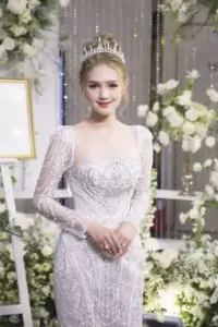 Cô gái tóc vàng diện váy cưới trắng ôm dáng, mỉm cười nhẹ giữa không gian hoa sang trọng