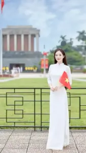 Nữ sinh áo dài trắng đứng trước quảng trường, tay cầm sách đỏ và mái tóc thả mềm