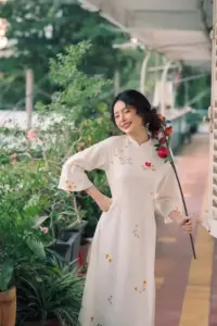 Cô gái mặc áo dài trắng họa tiết đứng trong vườn, tay vịn nhẹ và cười rất tươi