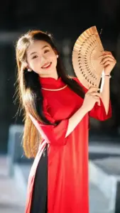 Nàng mặc áo dài đỏ cầm quạt tre, nụ cười rạng rỡ dưới ánh sáng ấm ngoài trời