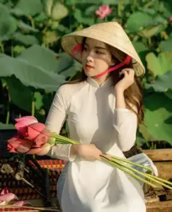 Cô gái áo dài trắng ngồi bên đầm sen, tay cầm cành hoa và dáng nghiêng mềm mại
