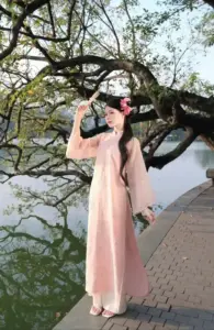 Thiếu nữ áo dài hồng đứng bên hồ, tay chỉnh tóc nhẹ dưới tán cây xanh mát