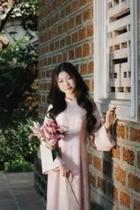 Thiếu nữ áo dài trắng cầm hoa bên tường gạch, tóc buông dài và nụ cười hiền