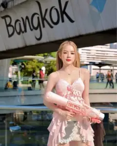 Phía trước bảng Bangkok, cô gái tóc vàng diện váy hoa hồng, ôm bó quà dễ mến