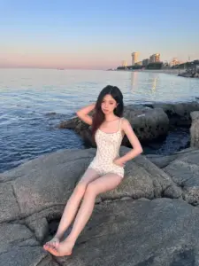 Trên ghềnh đá ven biển, cô nàng áo tắm trắng ngồi nghiêng, tóc xõa mềm nhìn rất dịu