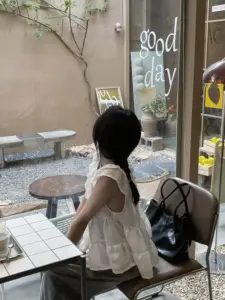 Thiếu nữ không lộ mặt ngồi cạnh cửa kính, áo trắng và túi tote tạo vẻ hiền