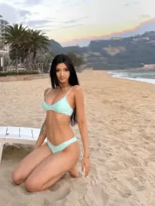Dưới hoàng hôn biển, bóng hồng TikTok bikini xanh ngọc quỳ trên cát, nét mặt dịu dàng