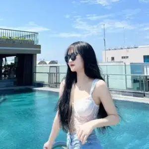 Cô gái đeo kính râm ngồi cạnh hồ bơi trong áo trắng tạo dáng thư thái dưới nắng