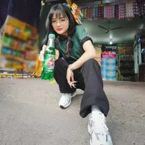 Cô gái áo xanh ngồi trong cửa hàng cầm chai nước với mái tóc đen và nụ cười tinh nghịch
